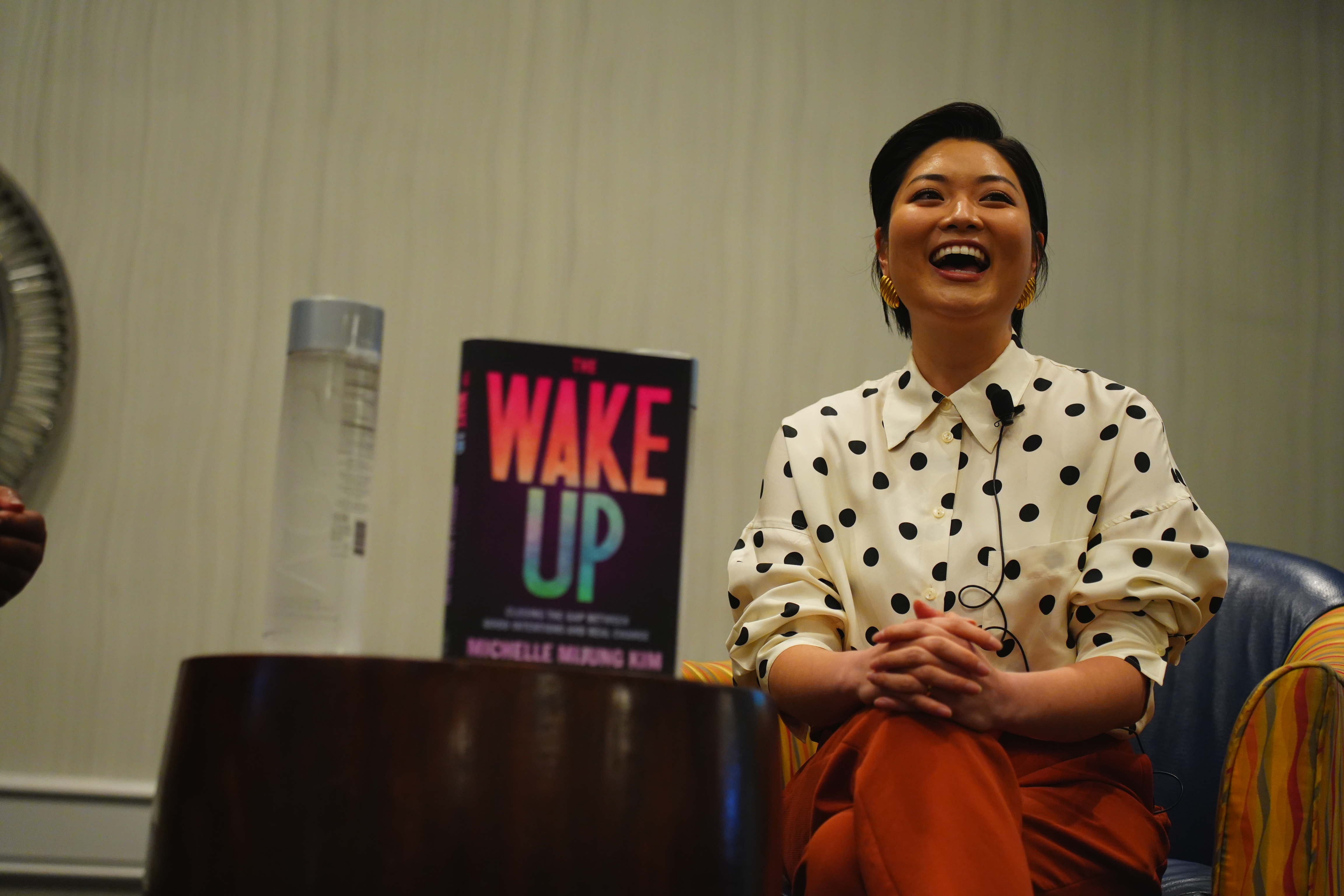 Michelle MiJung Kim laughing during a speaking event, with her book The Wake Up displayed beside her